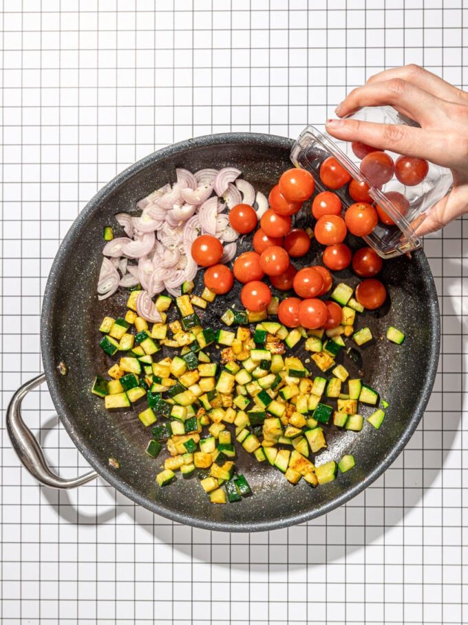 hand adding tomatoes to a skillet with zucchini