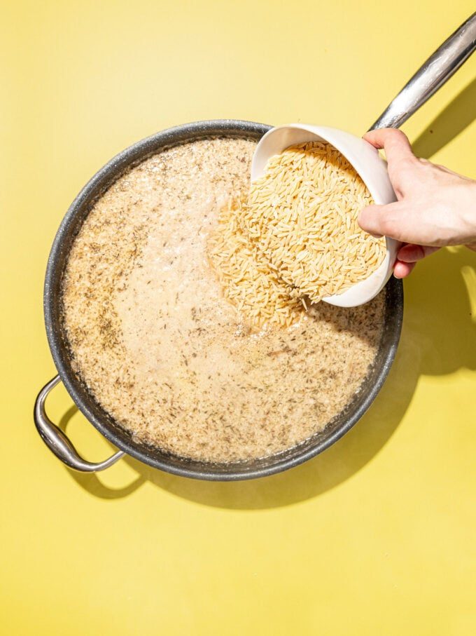 hand pouring orzo into skillet