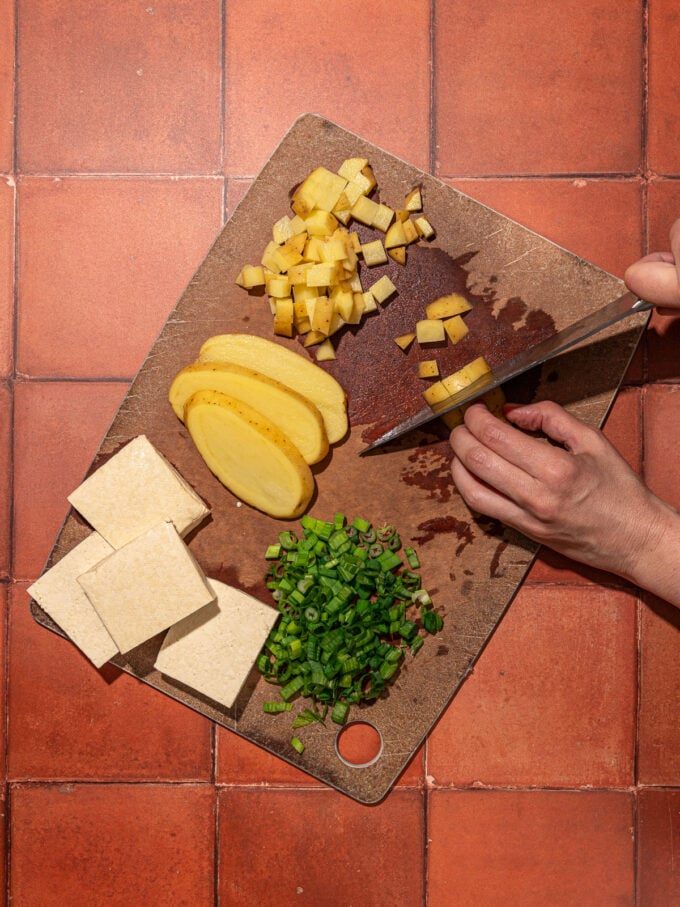 hand slicing potato on wood cutting board with green onion and tofu