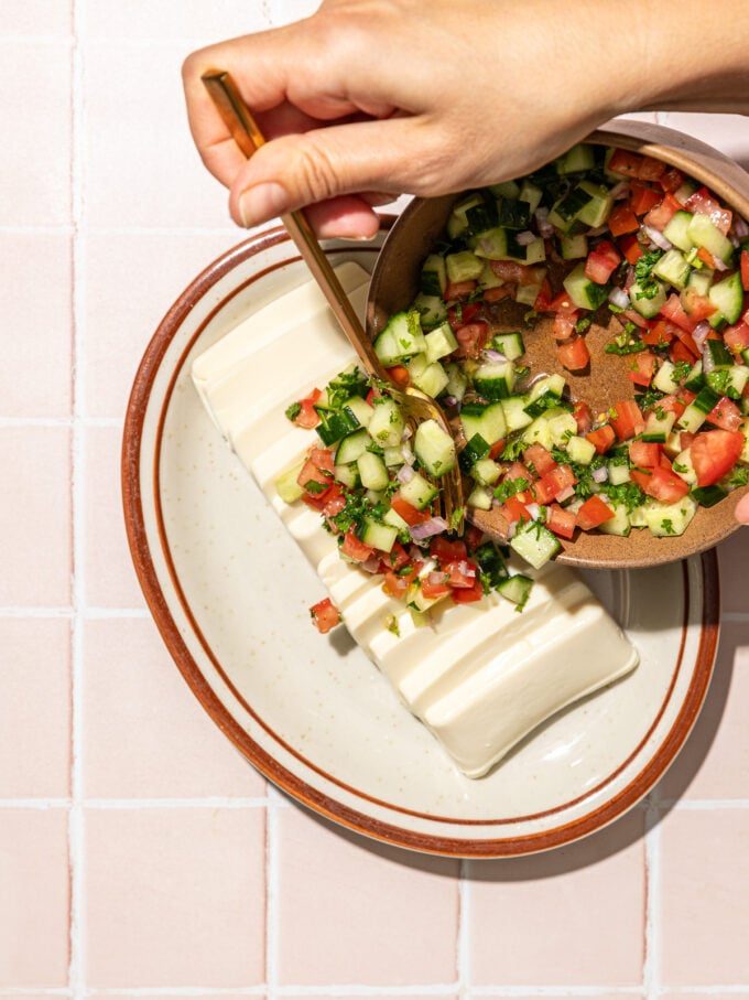 hand pouring salad onto sliced tofu on oblong plate