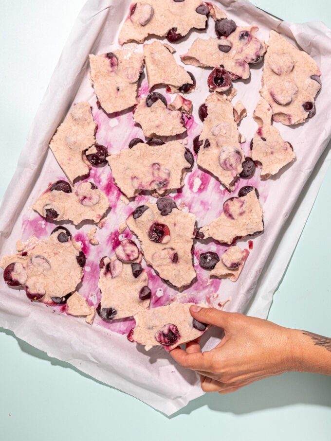 hand grabbing piece of cherry yogurt bark from baking sheet