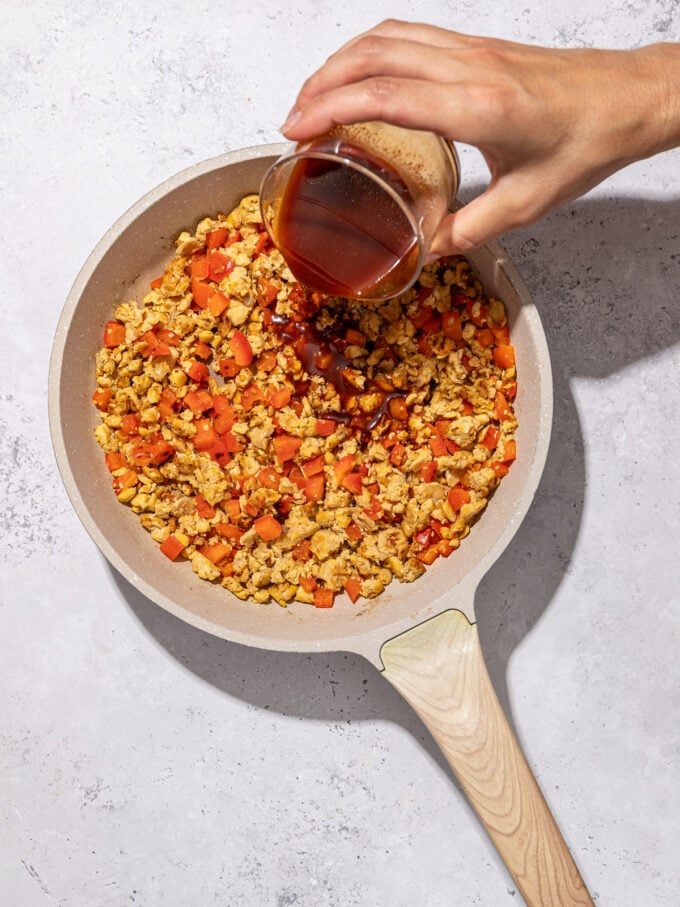 hand pouring sauce into skillet of tempeh and bell pepper